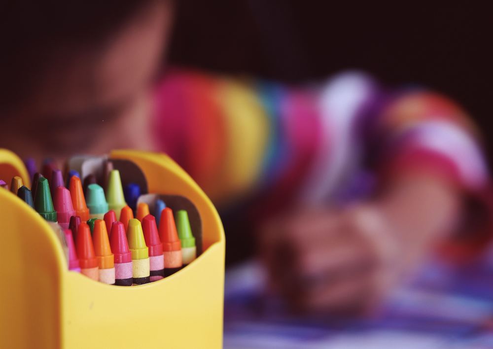 A boy colors in the background with the focus on a box of crayons in the foreground.